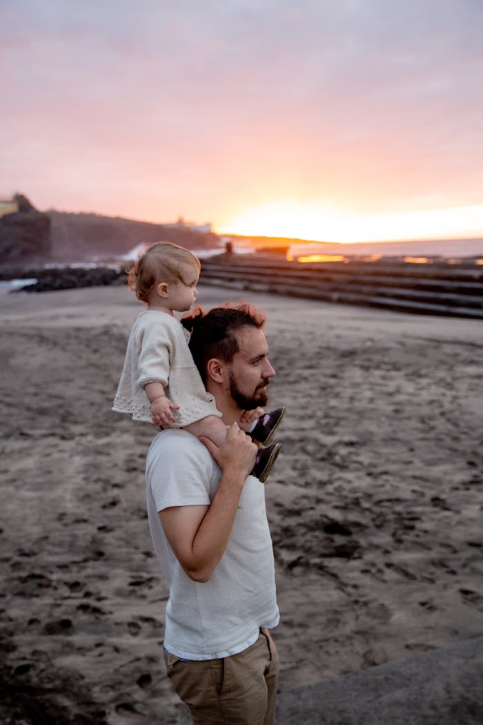 Father carries child on shoulders along a beach during a breathtaking sunset.